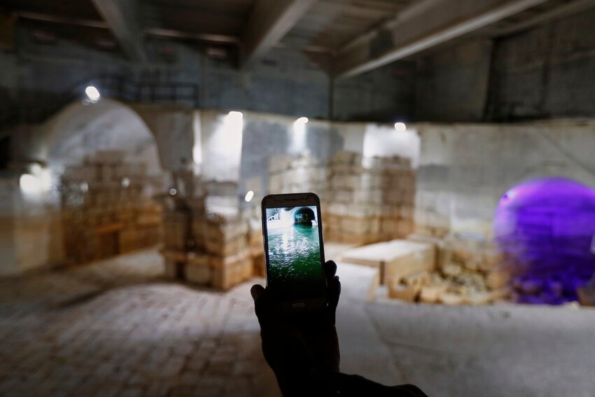 A worker holds up a cell phone displaying a photo of the Ein el-Fijeh reservoir, now seen dry in the background, in the Barada Valley, Syria, Thursday, May 8, 2025. (AP Photo/Omar Sanadiki)