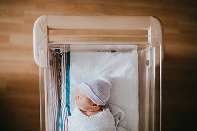 A baby just born at the hospital rests in a hospital bassinet crib, wrapped in a swaddle and wearing a beanie hat.