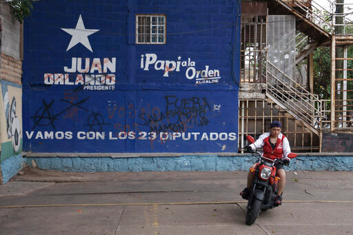 A woman rides her motorcycle past a campaign mural promoting the National Party presidential candidate Nasry Asfura known as "Papi a la orden",  in the Kennedy neighborhood of Tegucigalpa, Honduras, Friday, Nov. 26, 2021. Honduras will hold general election on Nov. 28. (AP Photo/Moises Castillo)