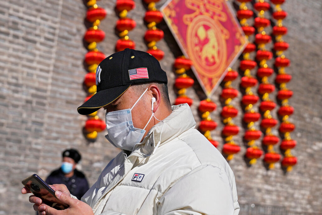 A man wearing a hat baring an American flag and face mask to help protect from the coronavirus walks by a masked security guard near lanterns decoration on the remnants of a city wall in Beijing, Thursday, Jan. 27, 2022. China is demanding the U.S. end “interference” in the Beijing Winter Olympics that begin next month, in an apparent reference to a diplomatic boycott imposed by Washington and its allies. (AP Photo/Andy Wong)