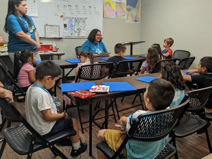 Charmaine Traffanstedt, sentada en el escritorio del maestro, con Laura Aponte, de pie a la izquierda, diseñó e impartió un campamento de ESL centrado en el Evangelio para niños de jardín de infantes a quinto grado en Cross Community Church en Houston.