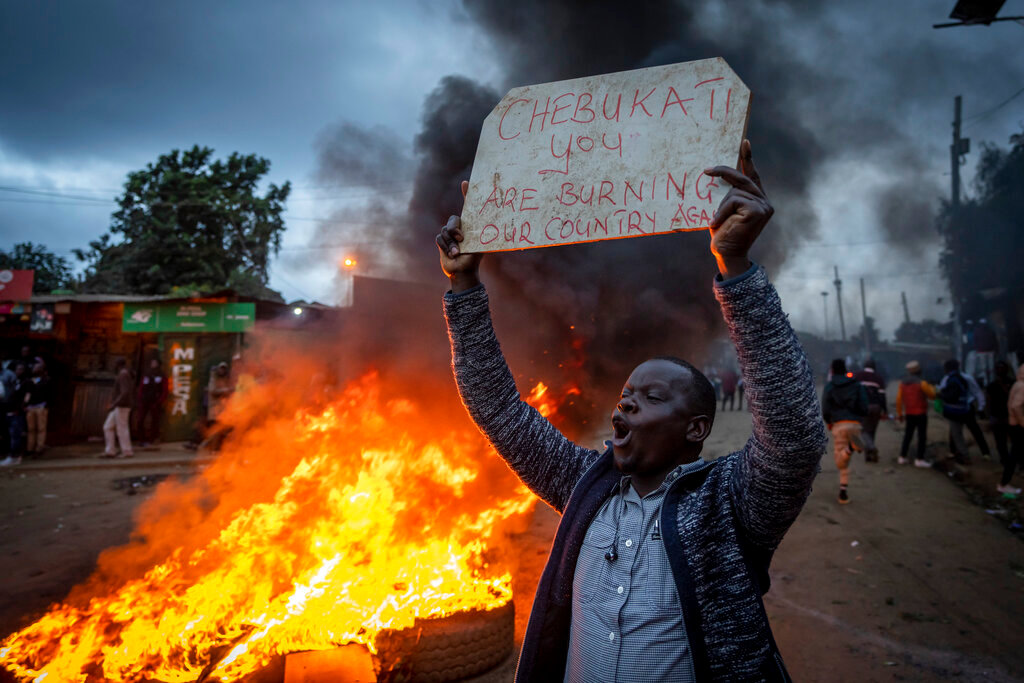 A supporter of presidential candidate Raila Odinga holds a placard referring to electoral commission chairman Wafula Chebukati, while shouting "No Raila, No Peace", next to a roadblock of burning tires in the Kibera neighborhood of Nairobi, Kenya Monday, Aug. 15, 2022. (AP Photo/Ben Curtis)