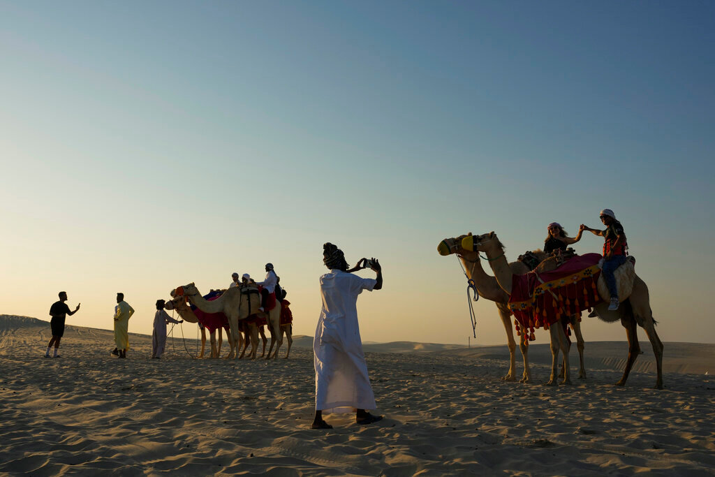 A guide takes a photo of a couple atop camels in Mesaieed, Qatar, Nov. 26, 2022. (AP Photo/Ashley Landis)