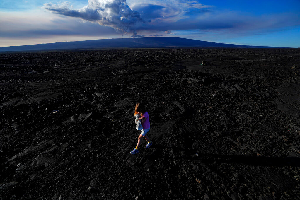 Abigail Dewar, of Alberta, Canada, holds a stuffed animal as she walks over hardened lava rock from a previous eruption as the Mauna Loa volcano erupts, behind, Wednesday, Nov. 30, 2022, near Hilo, Hawaii. (AP Photo/Gregory Bull)