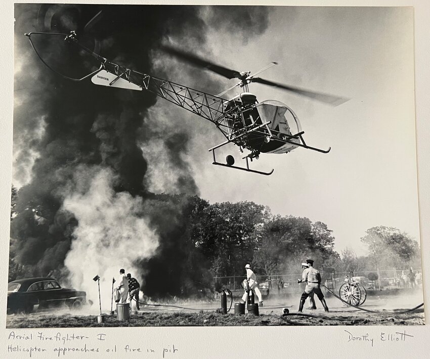 A black and white film photograph taken by former East Providence resident, Dorothy Elliott, in 1963 has a written inscription below: “Aerial firefighter…Helicopter approaches oil fire in pit.”