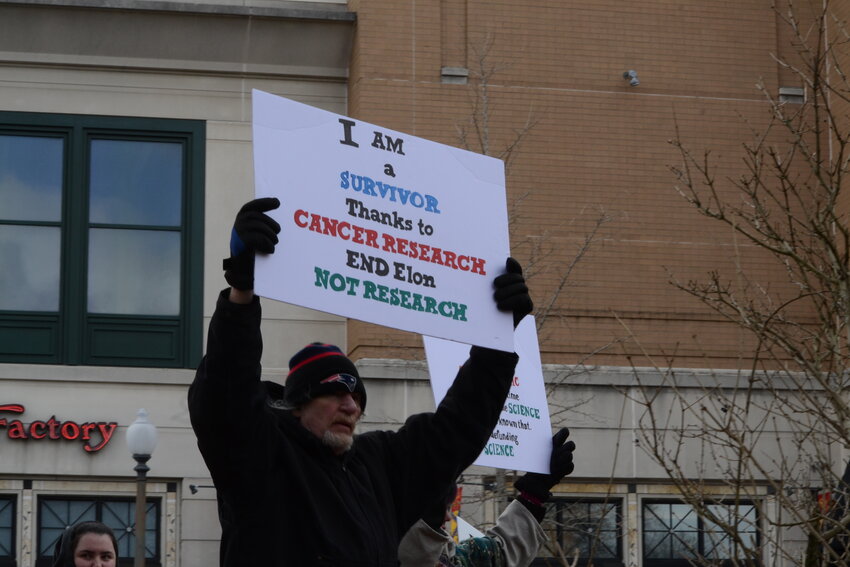A man holds a sign that reads “I am a survivor thanks to cancer research. End Elon. Not Research.”