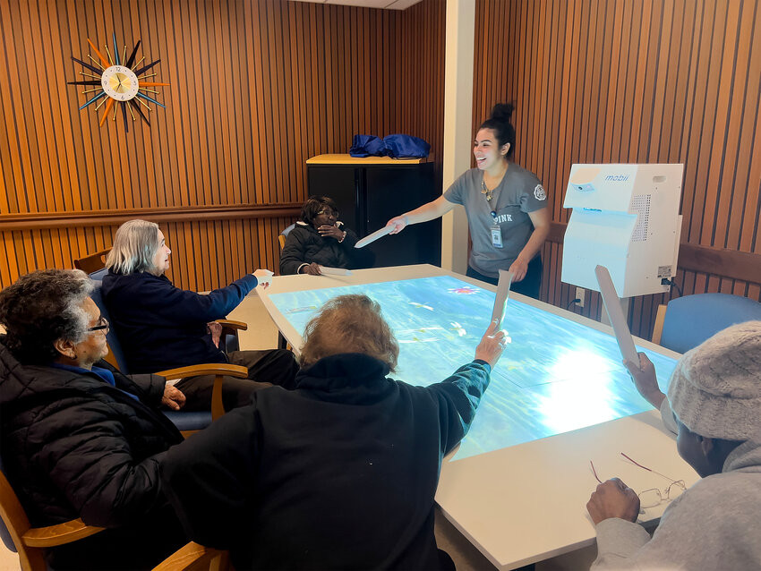 PACE daycare activities assistant Rosa Rivera (right) cheers on clients as they play a game of bopping the "Blue Suede Shoes" to Elvis' song during a game on the Mobii interactive table and floor system, which makes the participants move and have fun doing so. 