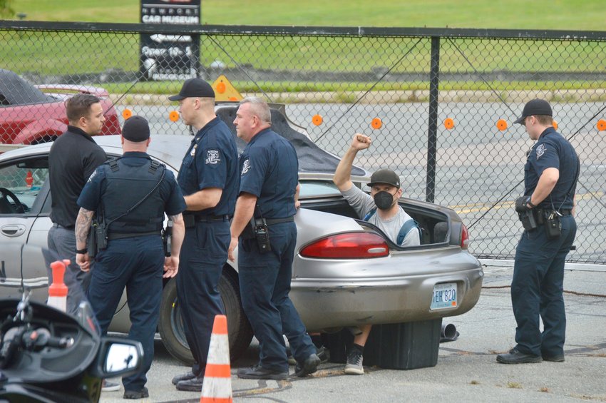 Protester Nicholas Katkevich, 36, of Bristol, who had cemented himself into the trunk of a car blocking the entrance to Raytheon Co., flashes a clenched fist as he&rsquo;s surrounded by Portsmouth police officers Thursday morning, Aug. 12. He was later extricated from the vehicle and arrested. Another protester was blocking the exit in a similar fashion.