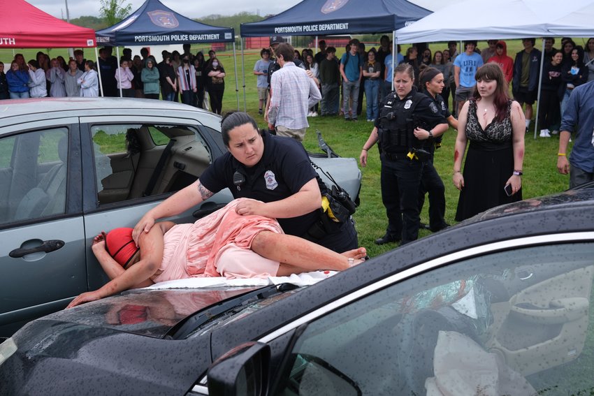 Police Officer Maddie Pirri checks Renesha Duncan for vitals during a mock DUI crash event staged outside Portsmouth High School in front of seniors last week. Duncan, a sophomore, was playing the part of a passenger who was killed after being ejected from a car operated by a drunk driver. Looking on at right is Anastacia Inman, another student who played one of Duncan&rsquo;s friends. In the middle are Police Officers Amanda Weaver (left) and JeanMarie Stewart.
