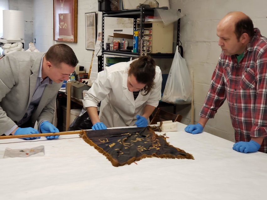 (L-r) Brendan Synnamon of the Union Drummer Boy Shop (a Civil War artifacts shop in Gettysburg, PA), Maria Vazquez of Royal Conservation Inc., and Ryan Meyer, Executive Director of the Naval War College Museum, inspect one of Bristol&rsquo;s two Babbitt Post 15 flags prior to restoration.