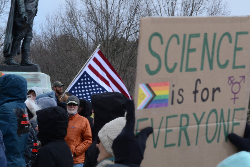 A protestor in the foreground holds up a sign that reads &ldquo;Science is for Everyone&rdquo; with the LGBTQ flag and the symbol that advocates for gender inclusivity.