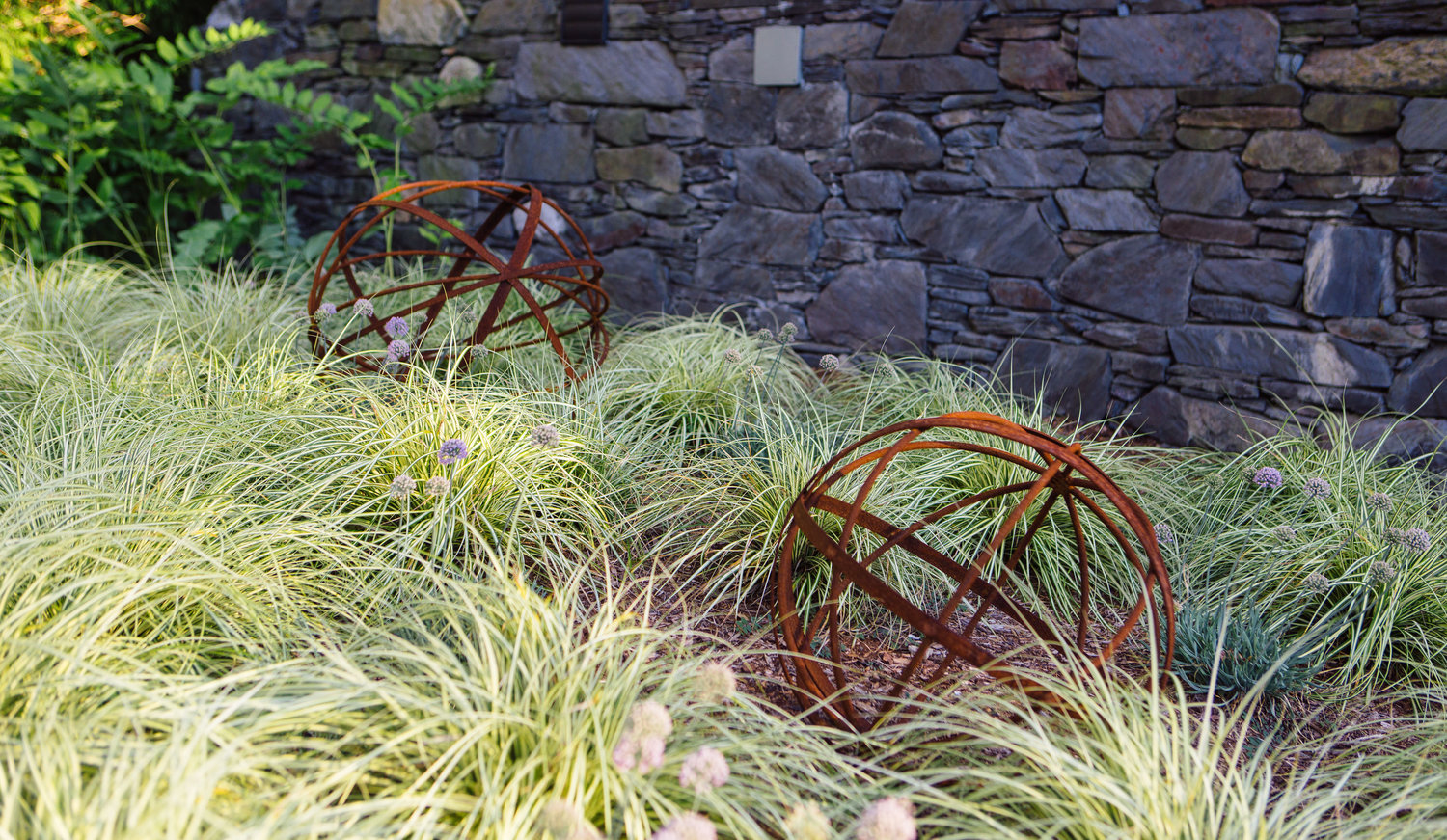 This small garden sits halfway up one of the flights of stairs, at a landing where a visitor sort of pauses on their way up. “I really felt it would be great to do something here,” Brook said. She found these metal sculptures and nested them amid an array of sedges and allium, creating an alluring little space. As seen throughout the property, it has that natural, wispy, relaxed feel about it.