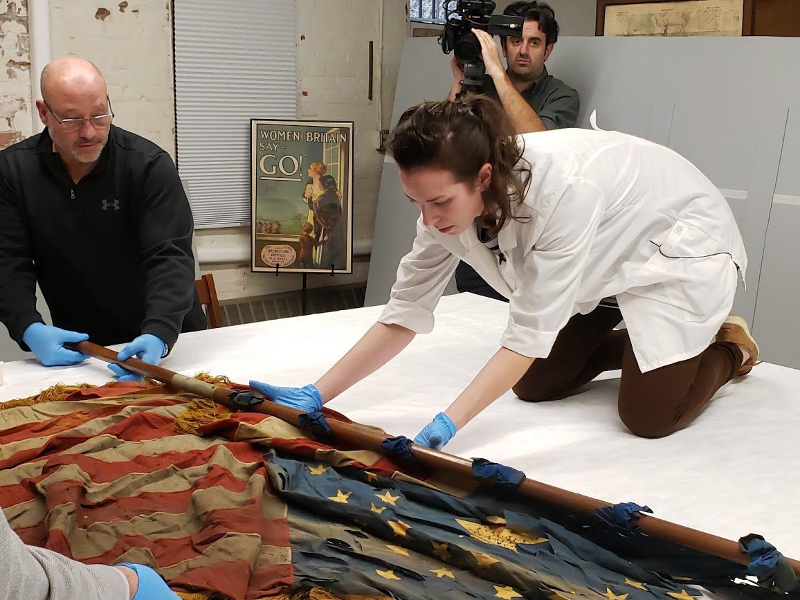 Expert textile conservator Maria Vazquez unfurls a U.S. flag from the mid-1870s, used by the local Bristol Grand Army of the Republic Post 15, known as the Babbitt Post. It was named for Bristol resident Jacob Babbitt of the 7th RI Volunteers, killed in action at the Battle of Fredericksburg on Dec 13, 1862. Andy Santilli, Varnum Armory contractor and volunteer, looks on.