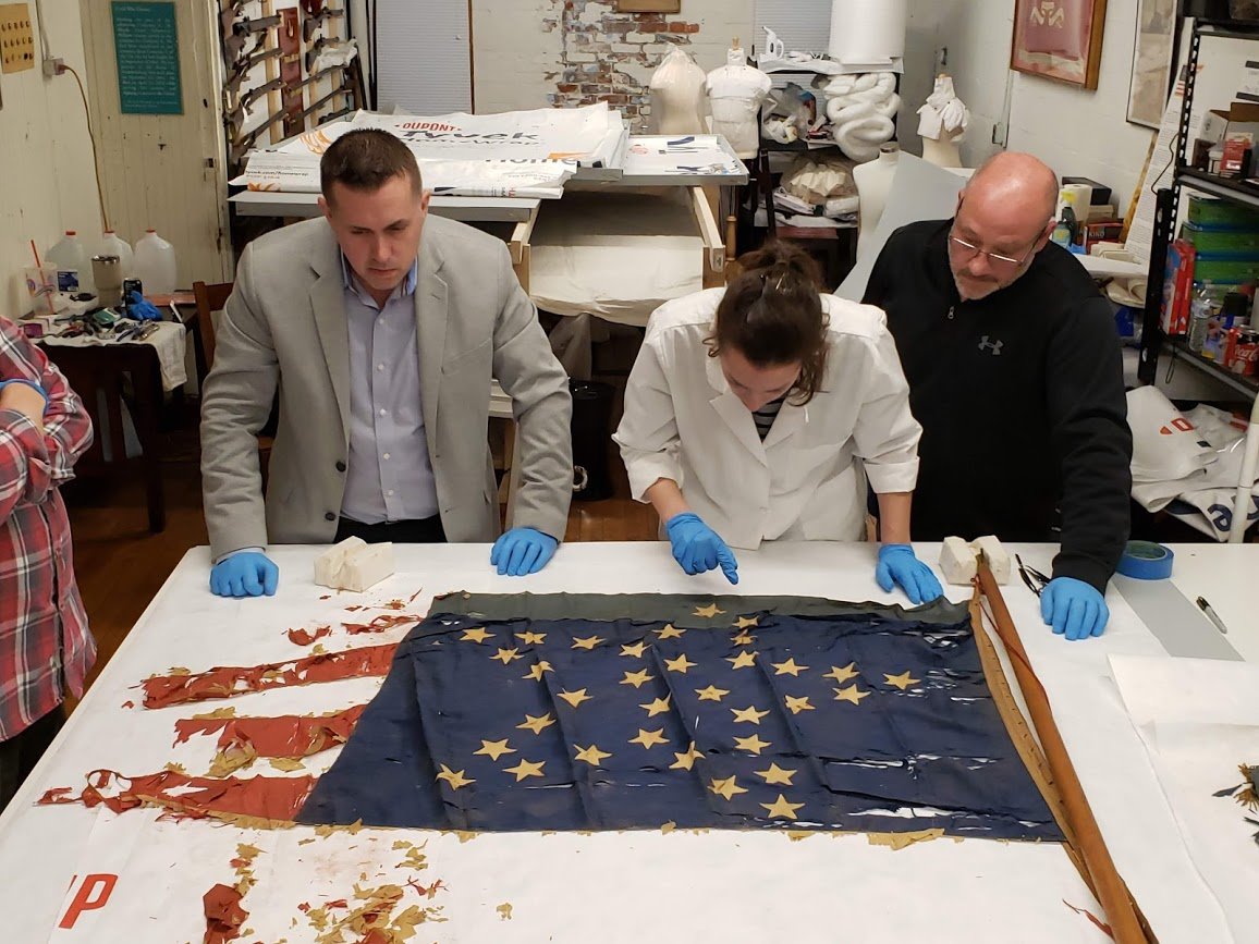 Brendan Synnamon, an expert appraiser and owner of the Union Drummer Boy in Gettysburg, Pa.; Maria Vazquez; and Andy Santilli, Varnum Armory contractor and volunteer, inspect the “Great Star” flag.