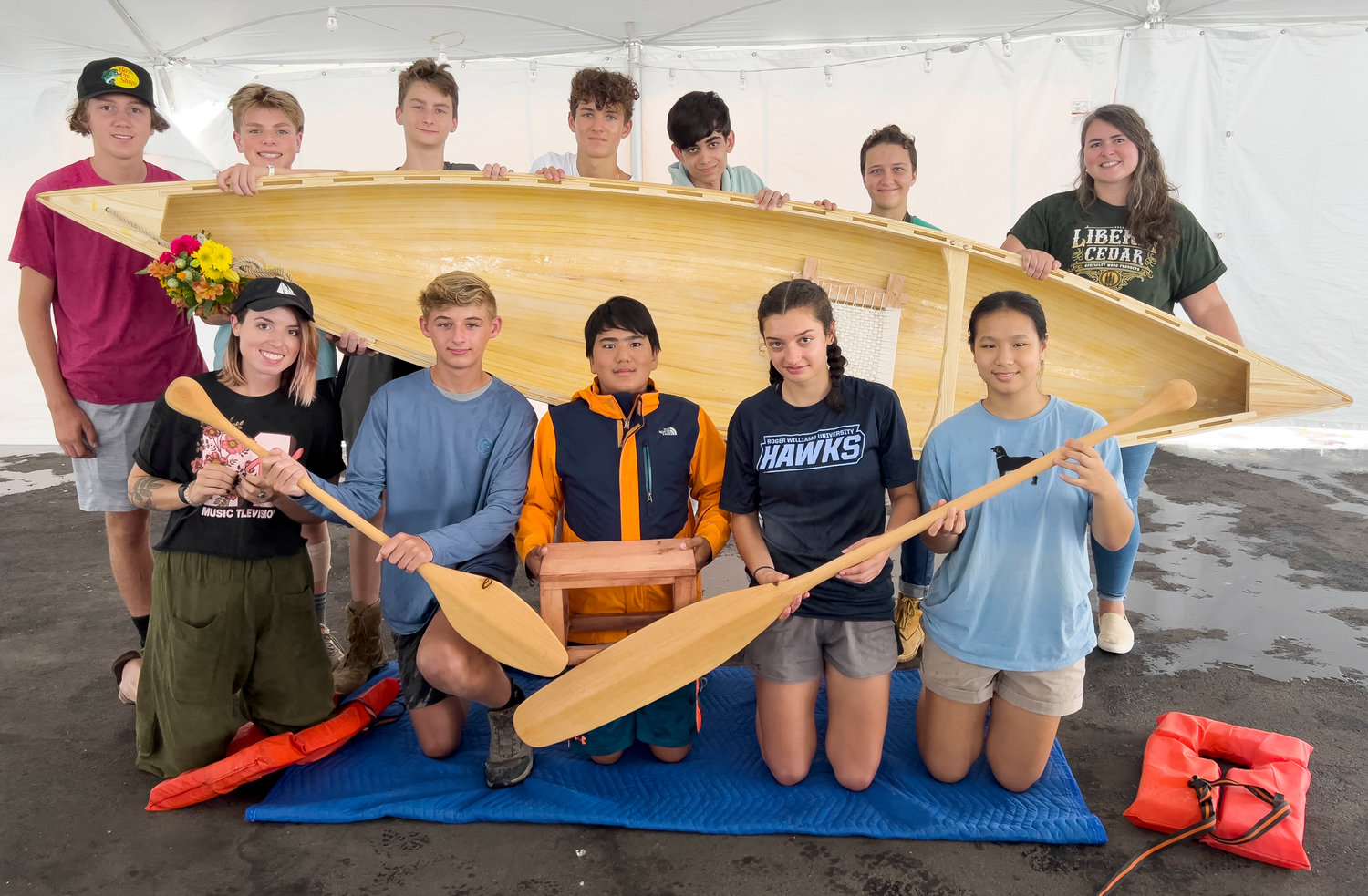 The boat building class poses with the one man wooden canoe that they built with paddles. Row one from left, assistant instructor Melissa Conlon,  Jarrett Rodrigues, Matthew Kibarian, Sonia Staroscik, Elena Sun, row two (top), Michael Kiselka, Colby Konz, Nickolas Markham, Erich Veegh, Aidan Enjeti, Thea Willner, instructor Ariana Murphy.