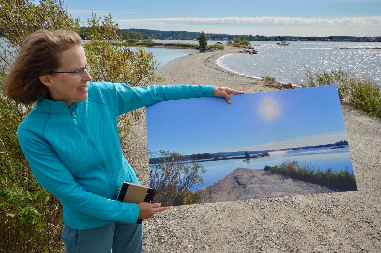 Save The Bay’s Wenley Ferguson holds a photograph, taken by a volunteer and shared on the MyCoast app, showing the access road (right behind her) to the former boat ramp at Gull Cove completely underwater during a king tide last year. “It shows how vulnerable this area is,” Ferguson said.