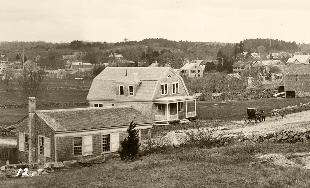 A view from Wolf Pit Hill looking west to Drift Road at the Head of Westport. In the far distance stands the Bell School and houses lining Drift Road. The Wolf Pit School, built in 1833, appears in the center foreground, close to the gambrel-roofed house built by W. F. King. On the far right stands the home of Dr. Tupper, who cared for seriously ill patients. Several buildings stand on the west landing across the river. Look closely to find a ladder leaning against a tree and the horse and buggy, probably belonging to the photographer. The photograph is from The Penobscot Marine Museum Collection, Eastern Illustrating and Publishing Company Collection.
