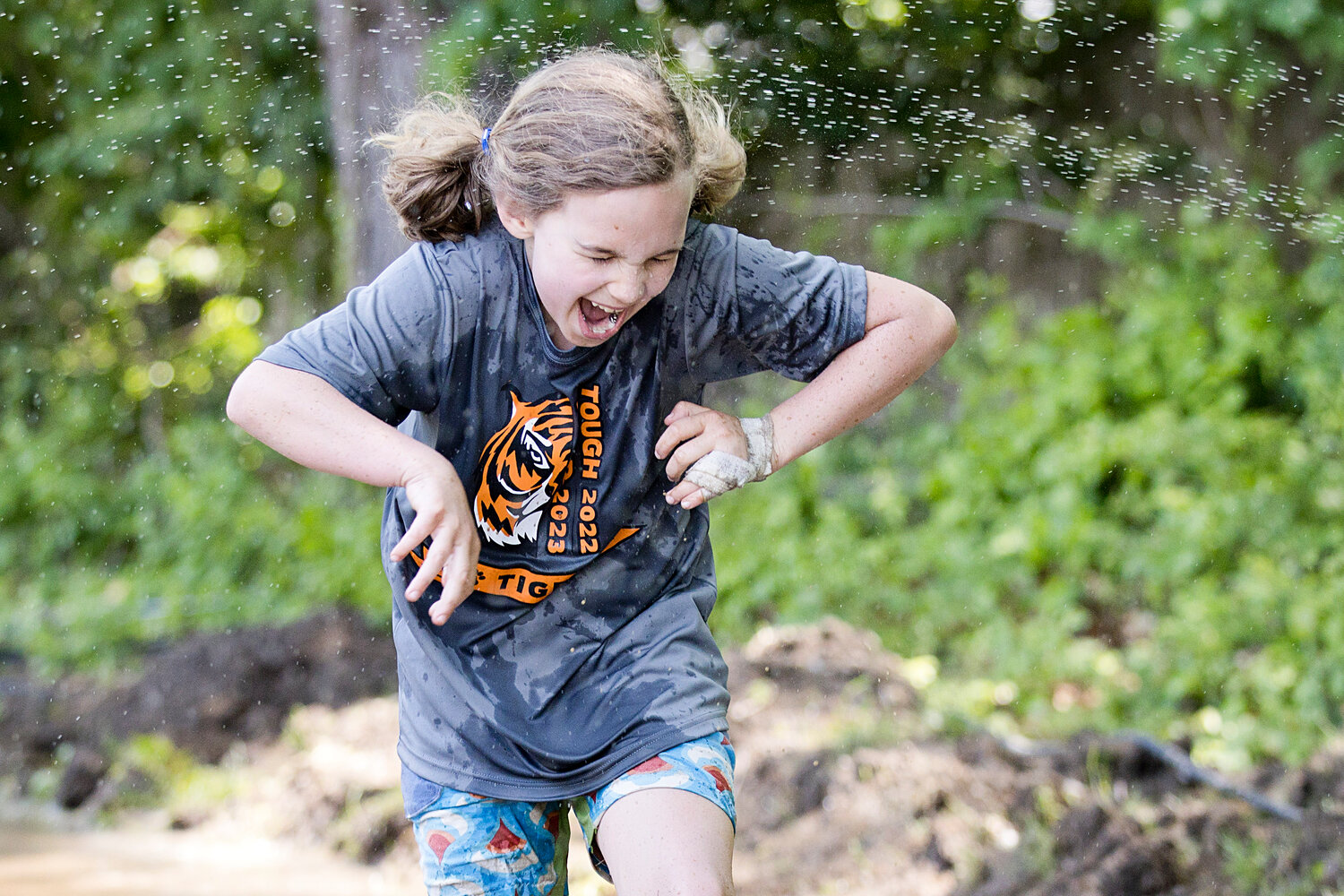 A young competitor reacts to an unsuspected sprinkler while racing through a mud obstacle.