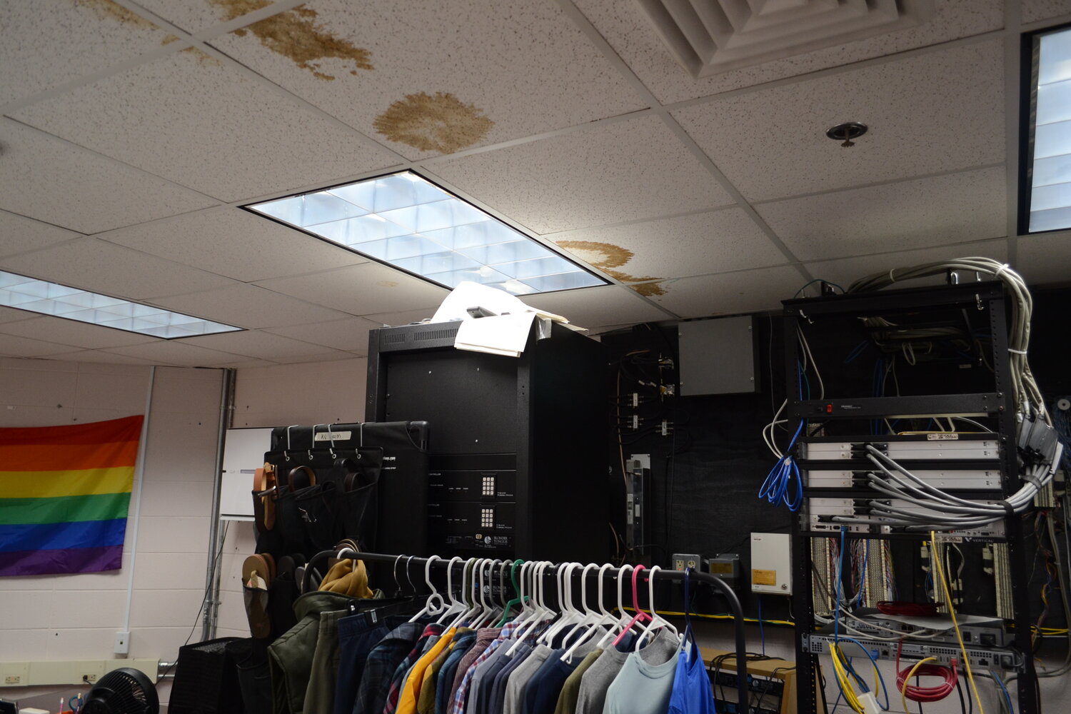 A stack of computer servers sits underneath visibly water damaged ceiling tiles in a room that also serves as a makeshift space for student volunteer work.