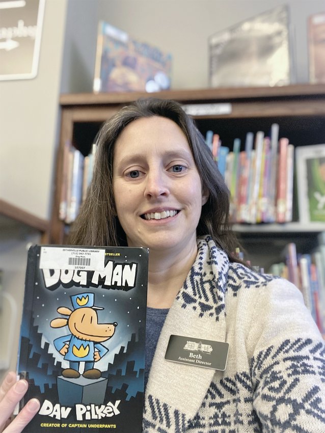 Beth Reineke, assistant director at the Estherville Public Library, shows off Dog Man, a selection from the library&rsquo;s children&rsquo;s section.   Photo by Amy H. Peterson