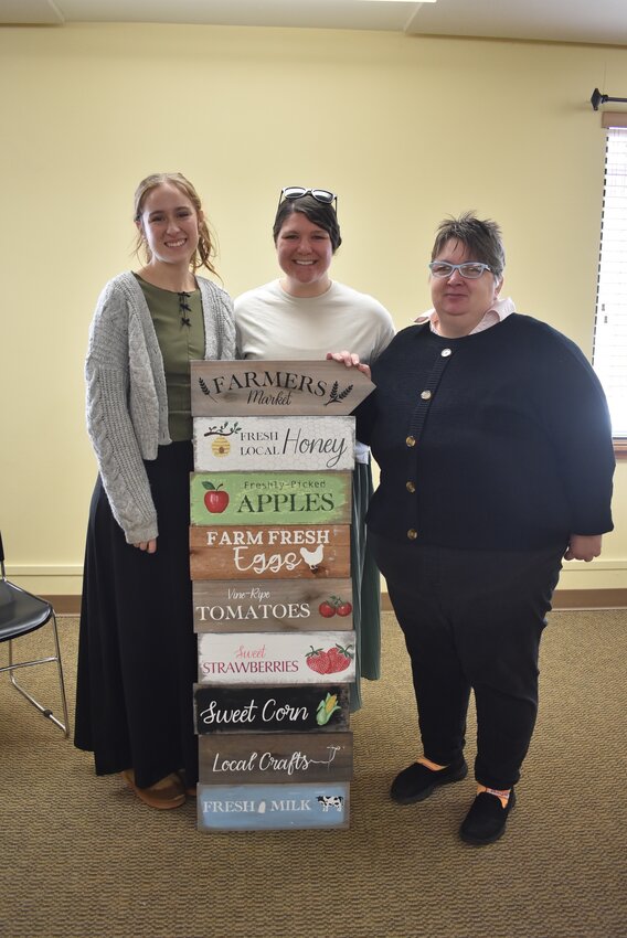 Angie Cain (right) poses with Tess Hillman (center) and Sarah Hoelzel (left) and one of the signs Hillman and Hoelzel have created for the new farmers’ market. Hillman and Hoelzel will be assisting Cain with marketing efforts and signage, through their printmaking company Living Designs.