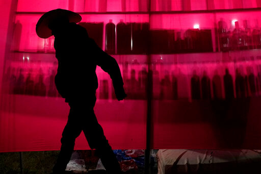 A man walks past liquor bottles silhouetted against a red cloth during the grand dance of the religious tradition, &quot;Folia do Divino Espirito Santo&quot; or Feast of the Divine, in the rural area of Pirenopolis, state of Goias, Brazil, Saturday, May 28, 2022. The event celebrates the coming of the Holy Spirit to Jesus' apostles after his crucifixion and has been performed for two centuries since it was first brought to Brazil by Portuguese colonizers. Once there, it was influenced by the cultures of Indigenous people and Black slaves. (AP Photo/Eraldo Peres)