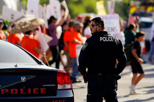 FILE - A Uvalde police officer watches as family and friends of those killed and injured in the school shootings at Robb Elementary take part in a protest march and rally, Sunday, July 10, 2022, in Uvalde, Texas.   Four months after the Robb Elementary School shooting, the Uvalde school district on Friday, Oct. 7 pulled its entire embattled campus police force off the job following a wave of new outrage over the hiring of a former Texas state trooper who was part of the hesitant law enforcement response as a gunman killed 19 children and two teachers.(AP Photo/Eric Gay)