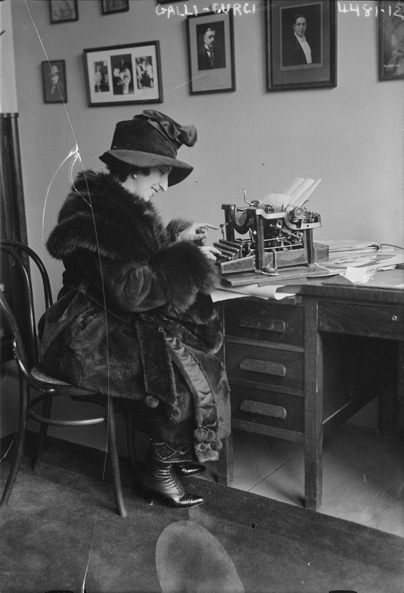 Famed Italian coloratura soprano Amelita Galli-Curci seated at a desk using a typewriter, dressed in fur coat and hat.