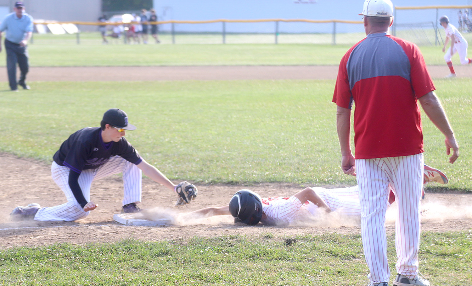 Fort Madison's Aiden Mitchell slides back into third safely after getting caught leaning on a pick-off attempt as Head Coach Ron Walker looks on from the third base coach box. The Hounds swept Keokuk 11-1, 5-1 in a doubleheader Monday night. Photo by Chuck Vandenberg/PCC