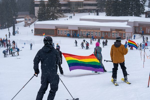 Skiers and snowboarders ride down the mountain with the pride flag.