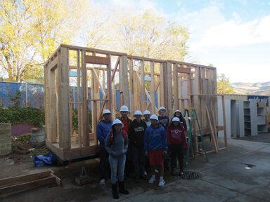 Students stand next to the framing as the walls are framed, upright, and ready for the next stage of building.
