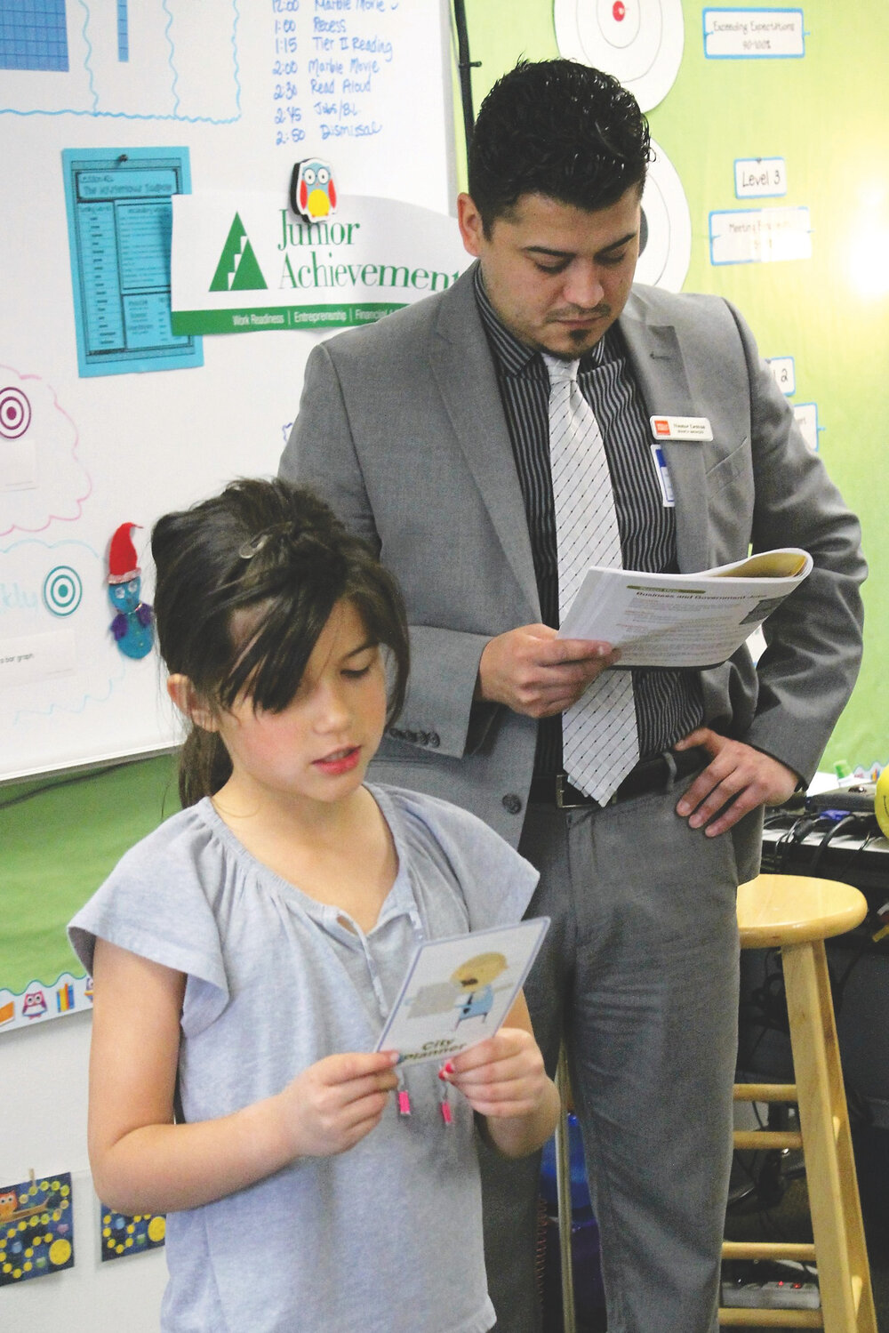 Brewster second grade student, Natalee Vandelac, reads a job description during the Teach Children to Save Day program presented by the Brewster branch of Wells Fargo Bank. Standing behind Vandelac is Wells Fargo branch manager, Nestor Lemus.