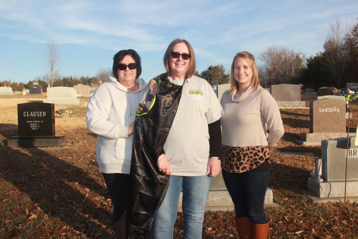 HIDDEN HERO — Andrea Romaker, center, works throughout the year to organize fundraisers and volunteer groups so that more than 550 wreaths can be laid at the graves of veterans at Christmastime. She was presented with her very own superhero cape by Mandy Andrews of the Warren County Record, while doing prep work at the Holy Rosary Cemetery with fellow volunteer Donna Mitchell, left.