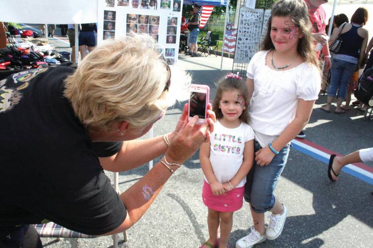 IN FULL BLOOM: Artist Toni Andersen of Wow Face Art snaps a shot of her handiwork on the faces of Hannah Fidas and her younger sister Alexis to post on Facebook. The Andersens and Fidas are neighbors and regulars at the festival.