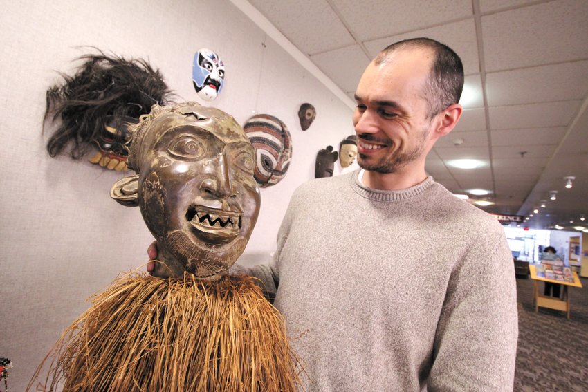 FACE TO FACE: Justin Bibee with a Bamum mask from Cameroon, specifically the Cameroon Grasslands, made of bronze with raffia palms. This mask is part of a family of masks that perform together. It represents the male human face and symbolizes a clan ancestor. Masks such as these are performed at funeral or memorial celebrations. This mask is worn on the top of the head, while raffia palm conceals the performer&rsquo;s face, and a mesh veil covers the performer&rsquo;s body. It is one of many in his collection on display at the Warwick Public Library through May 31.