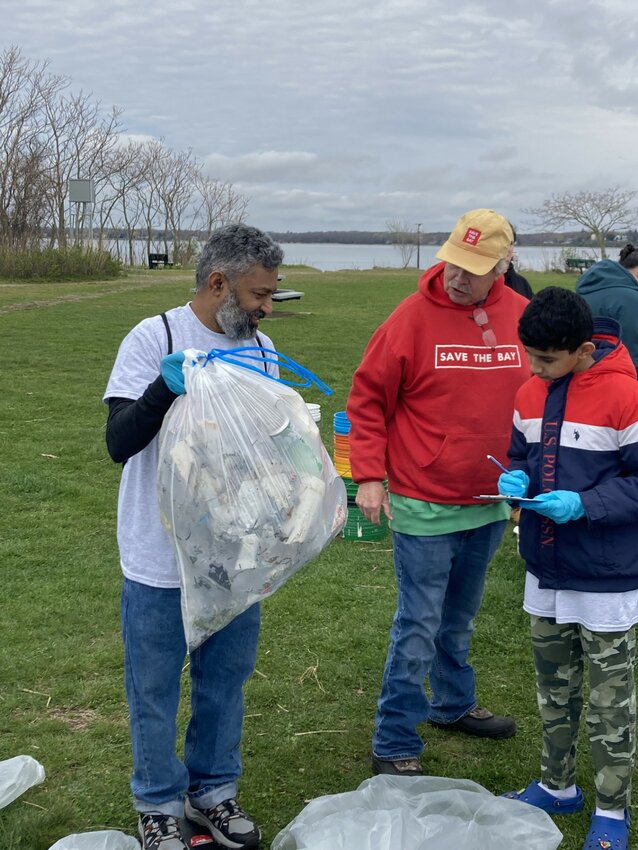 MARKING DOWN THE NUMBERS: Zakeer Shaik stands on a scale provided by John Paul, as the two talk about the trash they’ve picked up. Taking notes on how much each trash bag weighed is Zahid Shaik, Zaheer’s son. (Warwick Beacon photo)